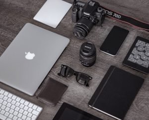 flat lay image of a macbook, canon camera, sunglasses and notepads